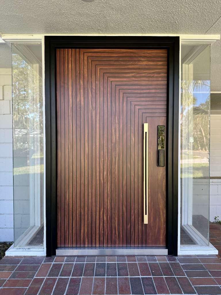 Wood-look pivot front door with a geometric grooved pattern, brass pull handle and smart lock in a black frame between glass sidelites