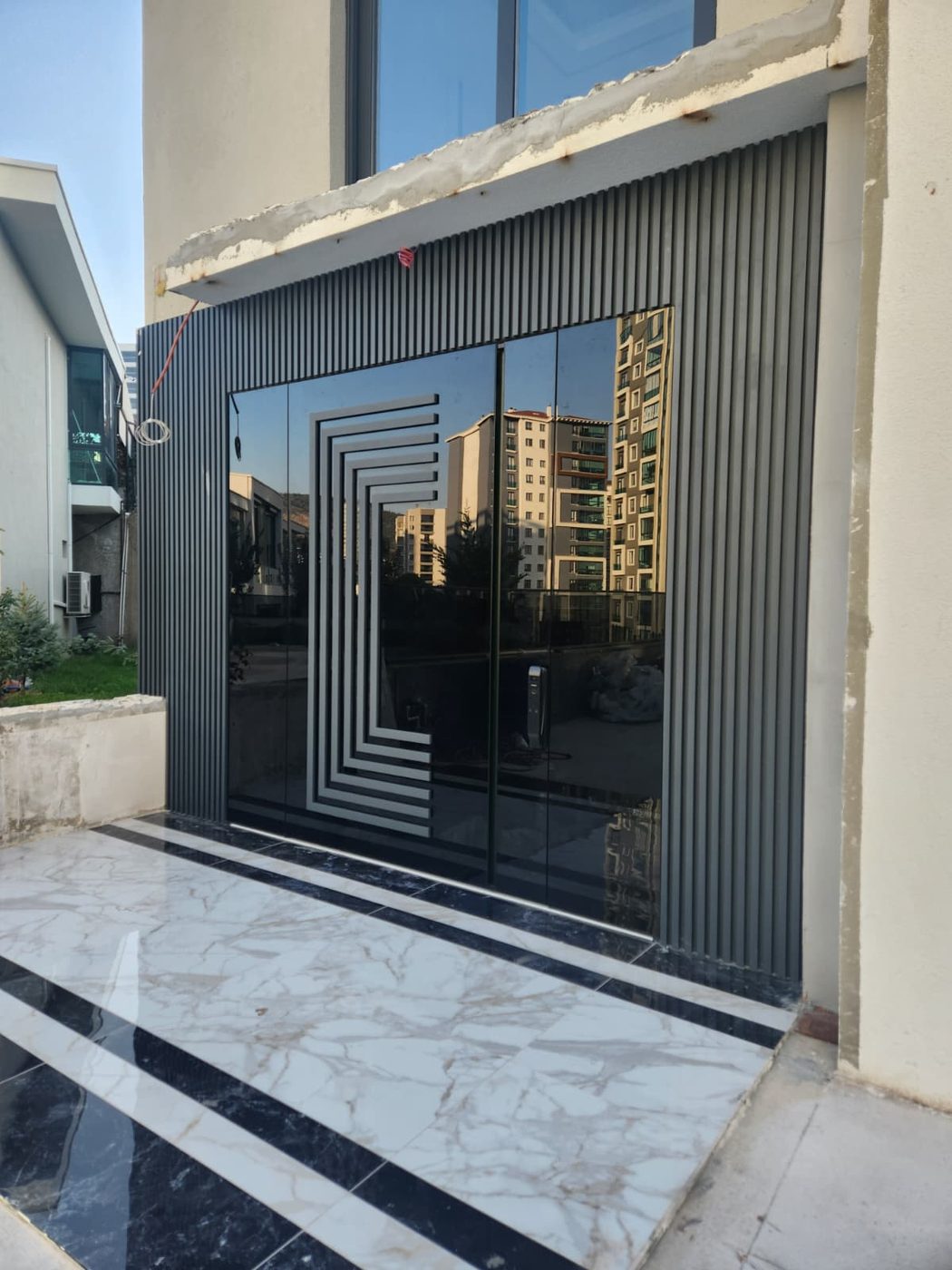 Black-glass double pivot entry with a stepped geometric inlay, framed by vertical aluminum slats
