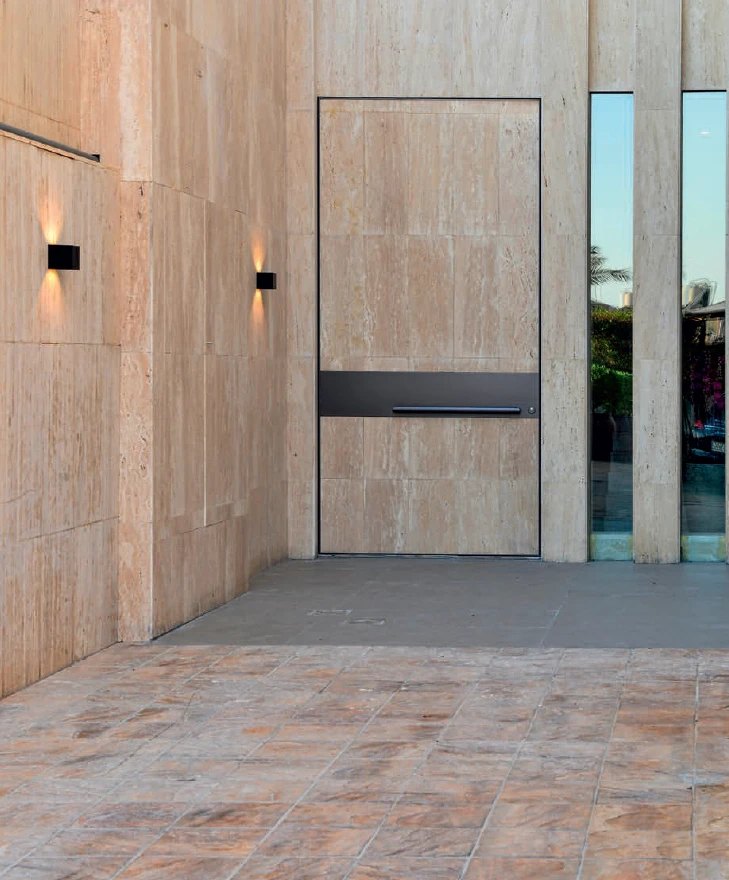 Travertine-clad pivot door with a bronze horizontal pull band in a travertine facade