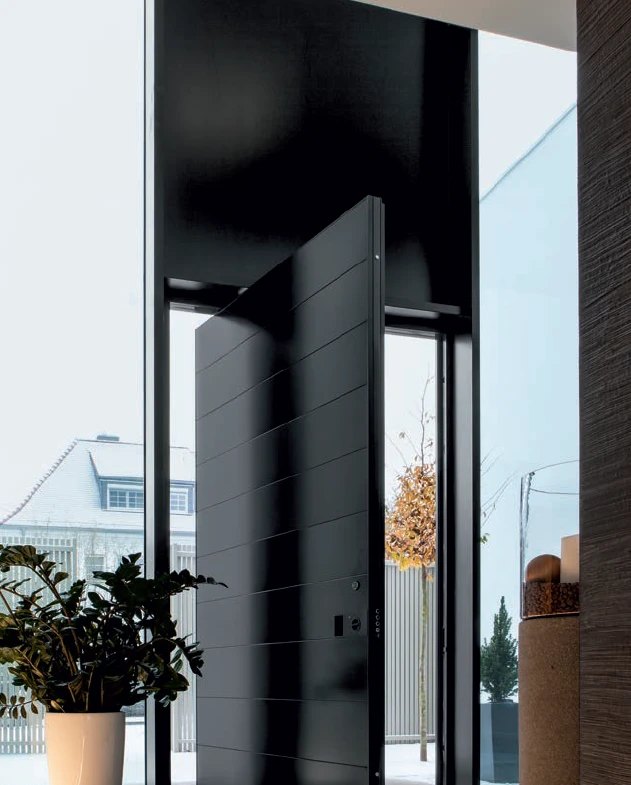 Double-height black pivot door with horizontal grooves, open in a tall glass entry